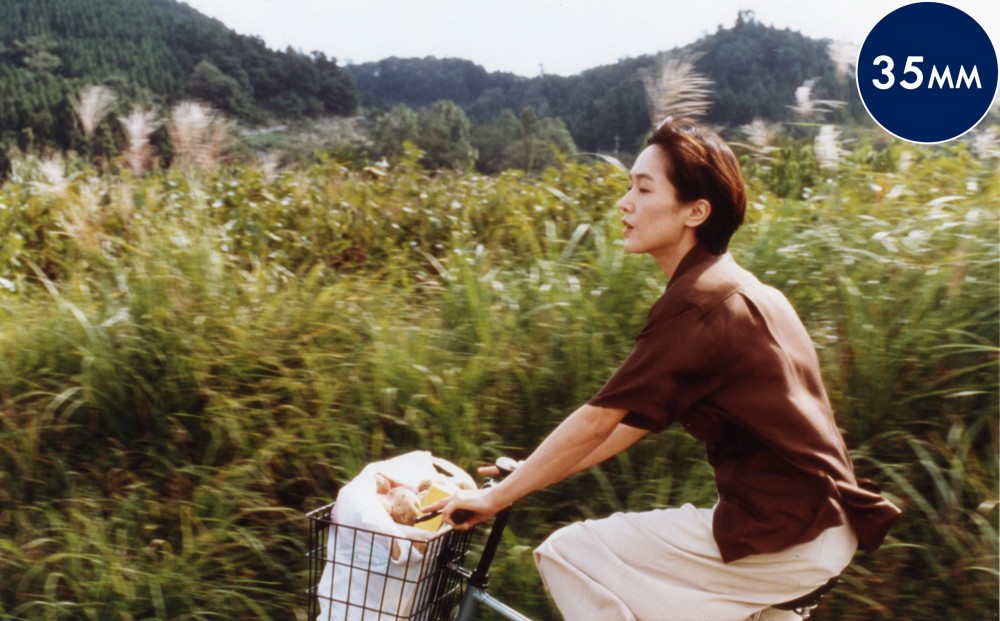 A woman rides a bicycle with tall green grasses and green hills in the background.