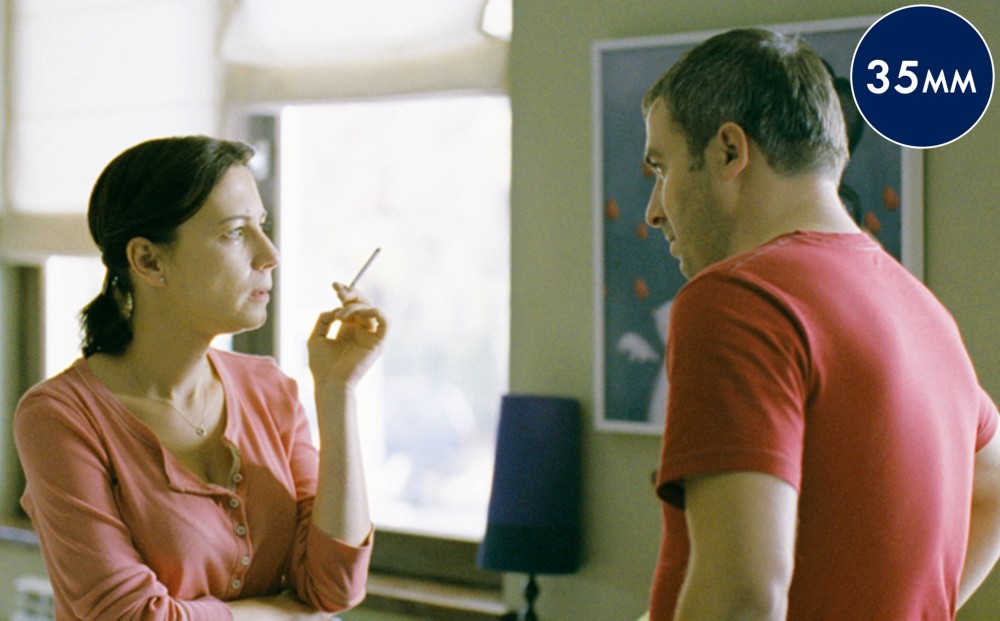 A man and woman look at each other, standing in a room; the woman holds a cigarette.