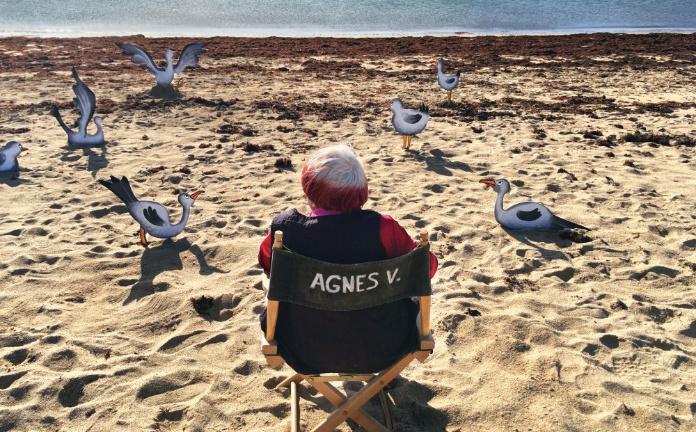 Director Agnès Varda sits in a director's chair on the beach, looking out at painted stand-outs of seagulls in the sand.