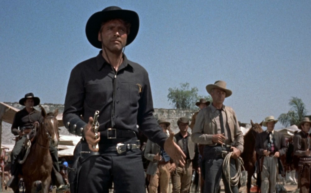 Actor Burt Lancaster brandishes his gun, a crowd of men behind - one on a horse, one holding a lasso, and others holding a gun.