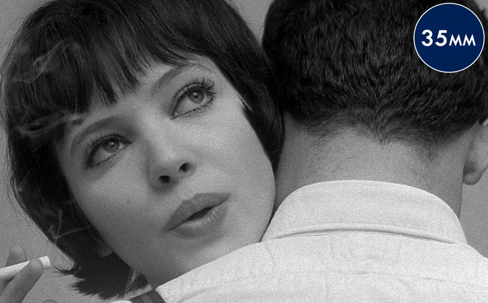 Close-up on Anna Karina's face, peeking over the shoulder of a man with his back to the camera.