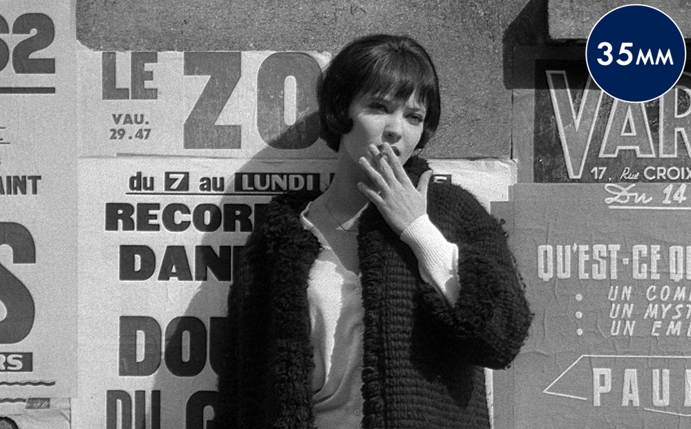 Actor Anna Karina smokes a cigarette and leans against a wall outside on the street.