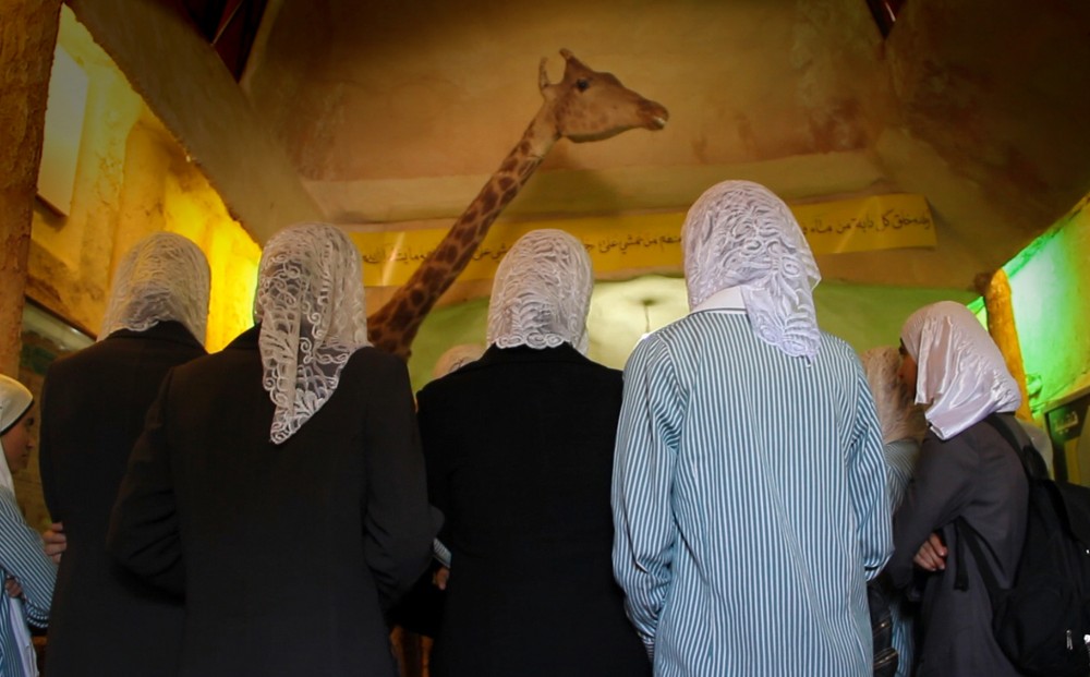 A group of people wearing hijabs stand near a giraffe statue, their backs to the camera.