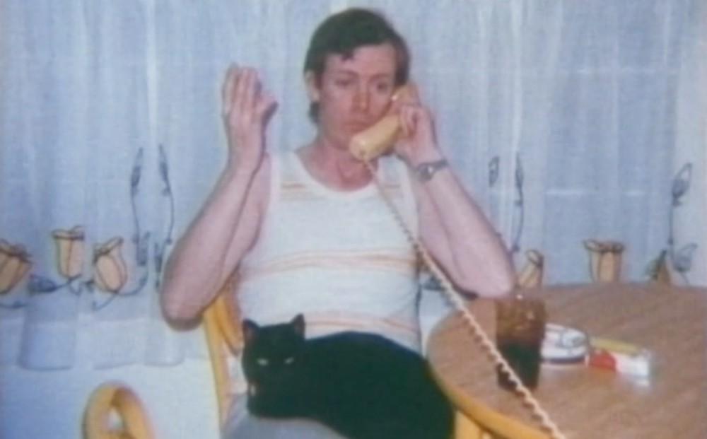 One of the film's subjects sits at a table, talking on a landline phone with a cat in their lap.