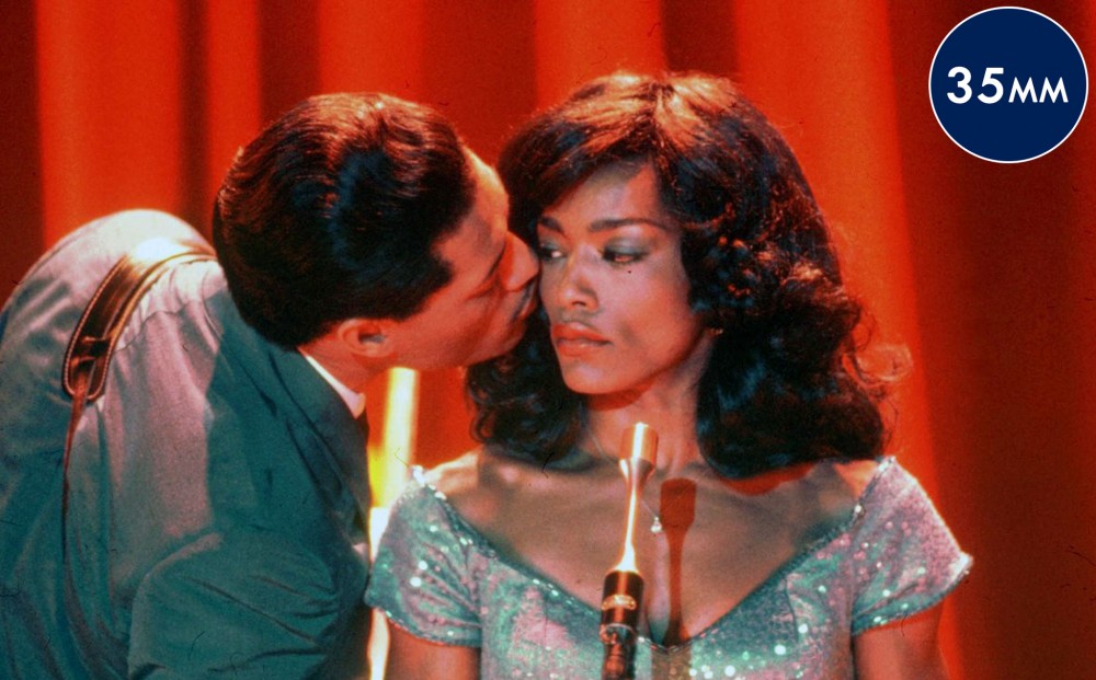 Actor Angela Bassett (as Tina Turner) stands in front of a microphone;  actor Laurence Fishburne (as Ike Turner) leans in, almost kissing her cheek.