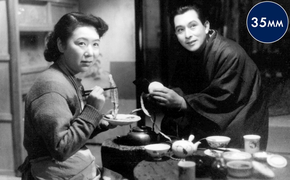 A woman and a man sit and eat together, both looking up at someone off-camera.
