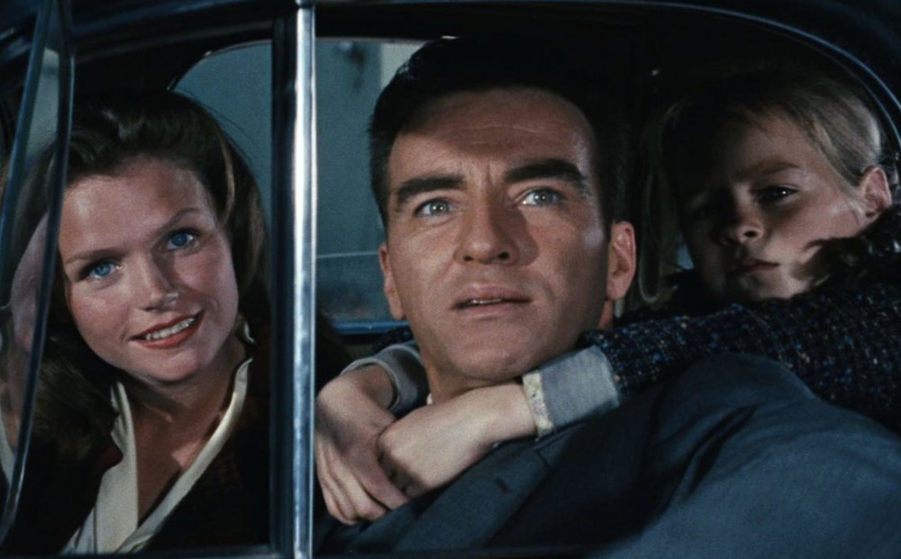 Actors Lee Remick and Montgomery Clift look out a car window; a small child sitting in the back seat has his arms around Clift's neck.