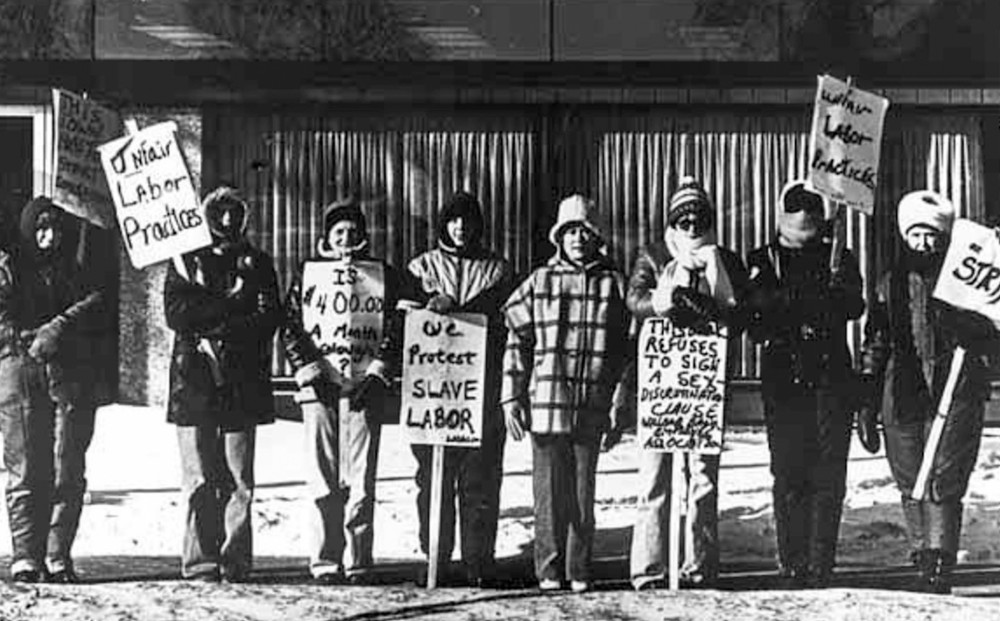 Black and white image of eight women standing in a row, holding picket signs.