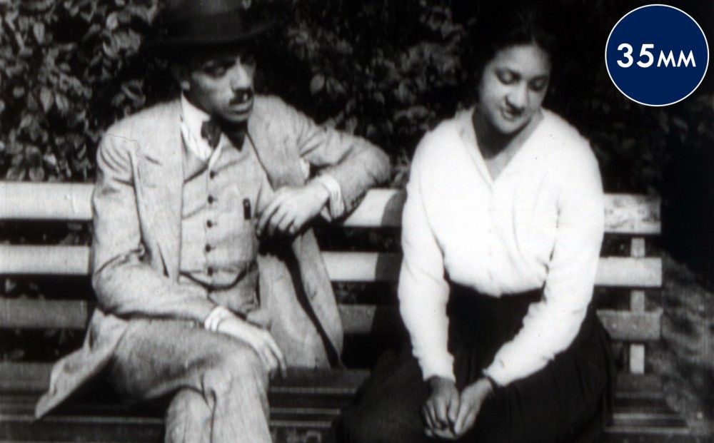 A man and woman sit next to each other on a bench; he looks at her and she looks away.