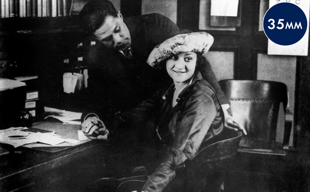 A woman sits at a desk; the man next to her holds her hand and rests his arm on the back of her chair. He looks at her, but she looks in another direction.