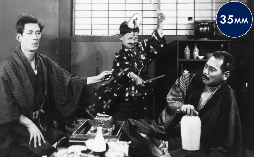 Two men sit with a young boy who wears a costume and and wields two knives.