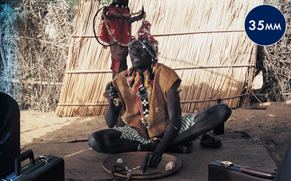 A man wearing a headdress with cowry shells sits on a rug in front of a thatched wall; briefcases are barely visible in the foreground.
