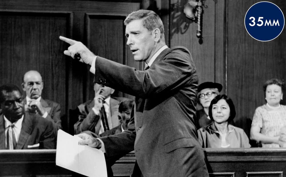 Actor Burt Lancaster stands in front of a jury box, holding papers and pointing enthusiastically.