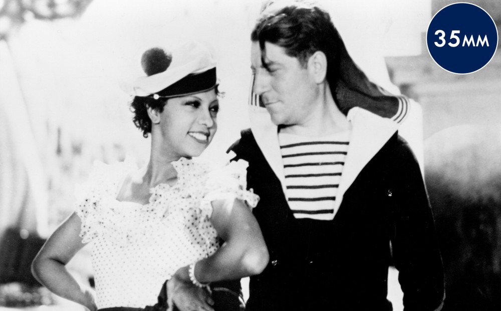 Actor Josephine Baker wears a sailor's hat and smiles; he stands next to her.