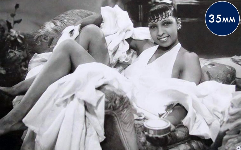 Actor Josephine Baker reclines in a chair and smiles, wearing a white dress.