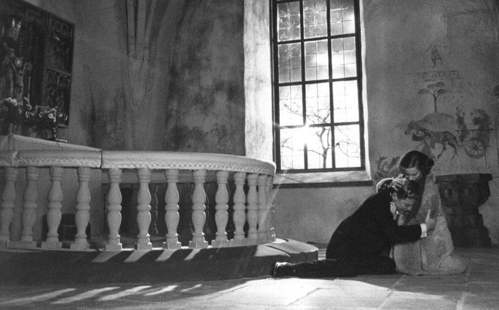 Actor Ingrid Thulin kneels beside and embraces Gunnar Björnstrand, who is sitting on the ground.